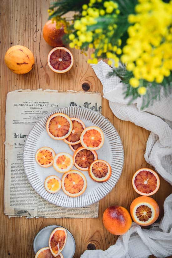 Des tranches d'oranges sanguines sont disposées sur une assiette, posée sur une table en bois. Un tissu, un journal ancien et du mimosa décorent la table.