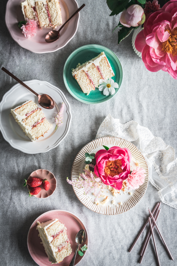 Vue de dessus d'une table de goûter. Un beau gâteau blanc à étages est découpé et les parts sont disposées dans de jolies assiettes aux couleurs printanières.